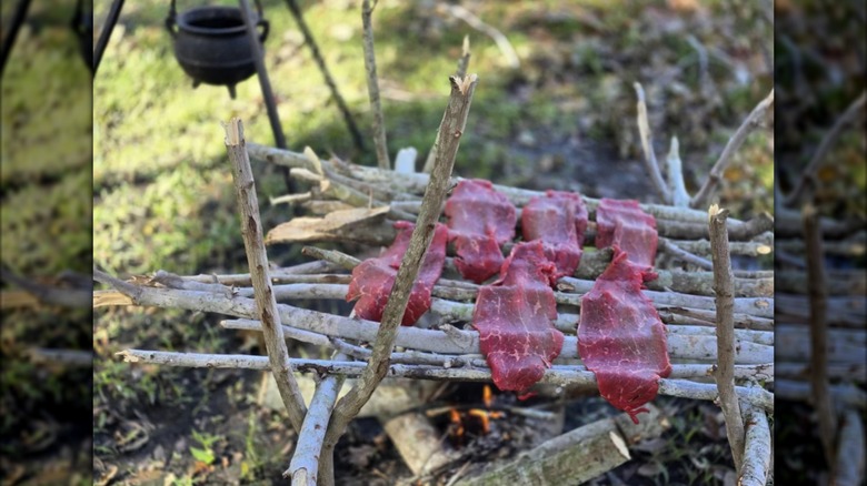 Raw meat being smoked/cooked on branches constructed into a grill above fire