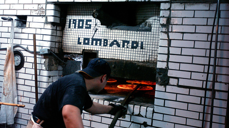 A pizza chef uses a peel to turn a pizza inside the original oven at Lombardi's, with the year 1905 displayed on its front