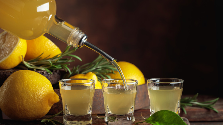 A bottle of Limoncello being poured into shot glasses with lemons nearby