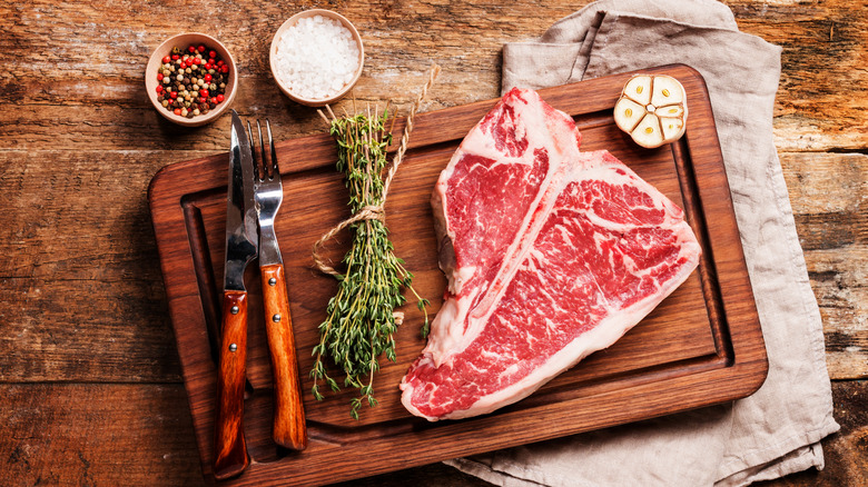 A raw porterhouse with a bundle of thyme sprigs, salt, multicolor peppercorns, and half a raw garlic bulb