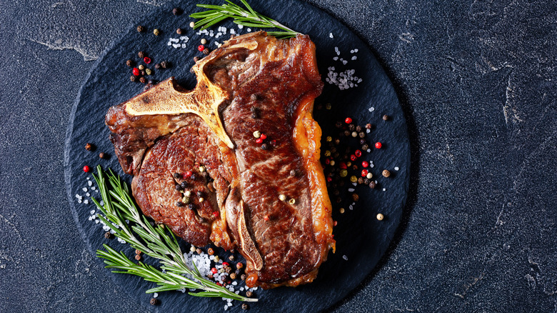 A grilled porterhouse on a black plate with salt, multicolor peppercorns, and rosemary sprigs