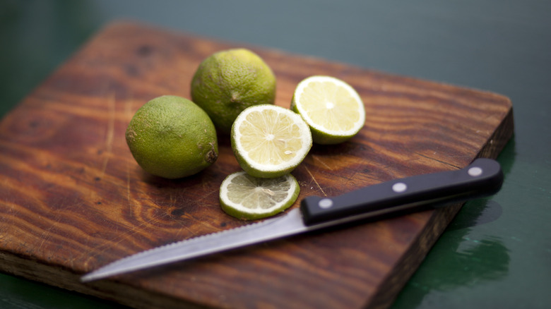 Close up of whole and cut limes on a wooden cutting board next to a knife.