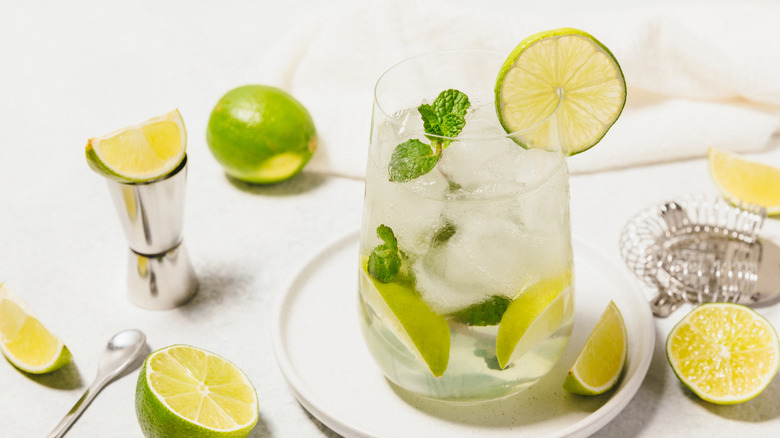Limes in a refreshing drink in a glass on a table surrounded by whole and cut limes.