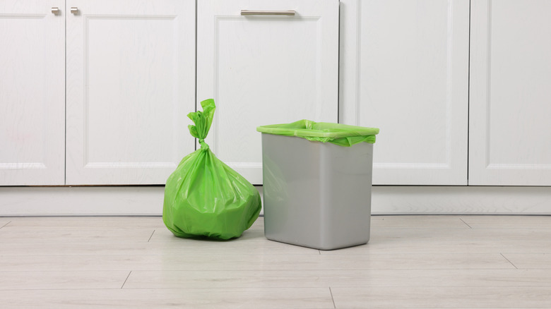 Full, green trash bag that's been tied off next to bin with a fresh bag in it in a kitchen with white shaker cabinets.