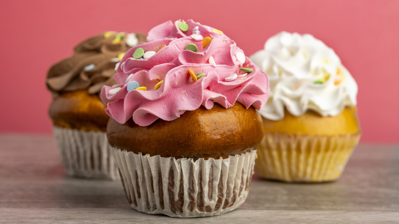 Three cupcakes each topped with either brown, pink, or white buttercream frosting, plus green, white, blue, and yellow sprinkles.