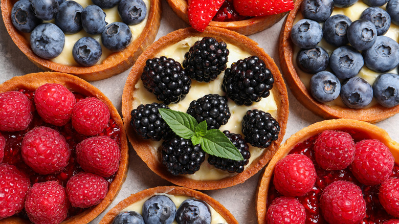 An assortment of fruit tarts, some filled with a berry jam and topped with raspberries. Others are custard with blueberries or blackberries.