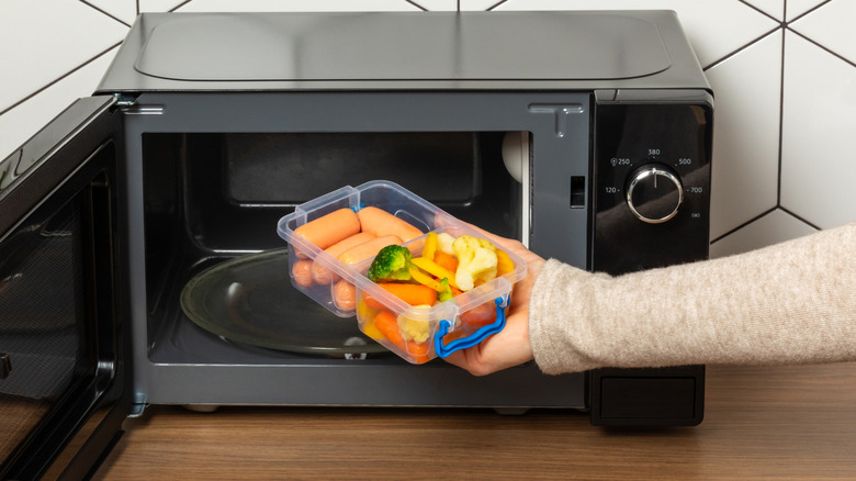 Hand placing plastic container full of vegetables in microwave