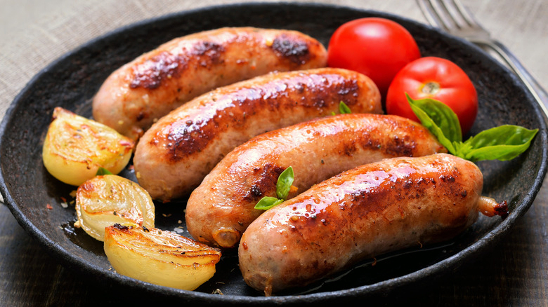 A pan of grilled bratwurst with roasted garlic cloves, two whole tiny tomatoes, and basil