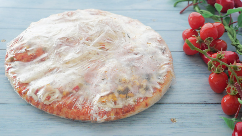 A wrapped frozen pizza on a table next to some cherry tomatoes on the vine