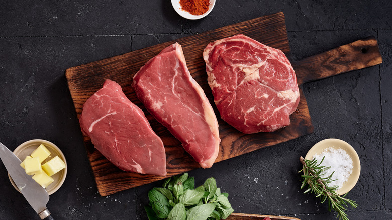 Different cuts of steak resting on cutting board with butter, salt, and herbs