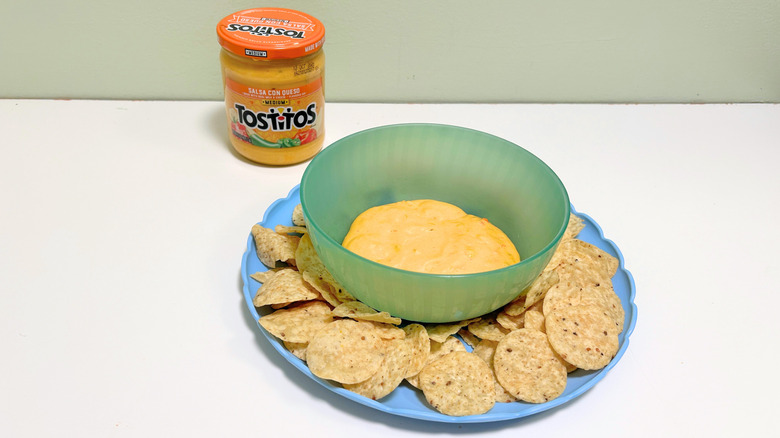 A bowl of queso surrounded by tortilla chips next to a jar of Tostitos Medium Salsa Con Queso