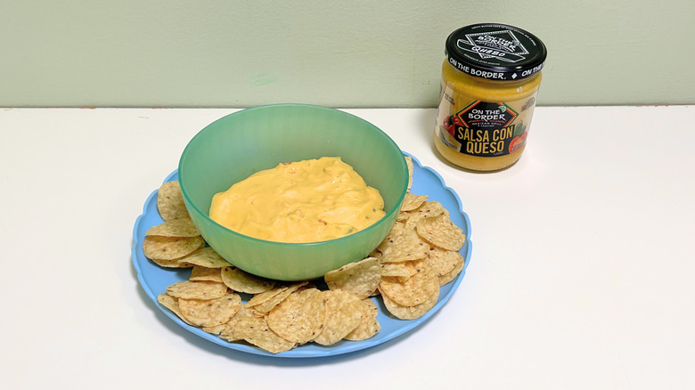 A bowl of queso surrounded by tortilla chips next to a jar of On the Border Salsa Con Queso