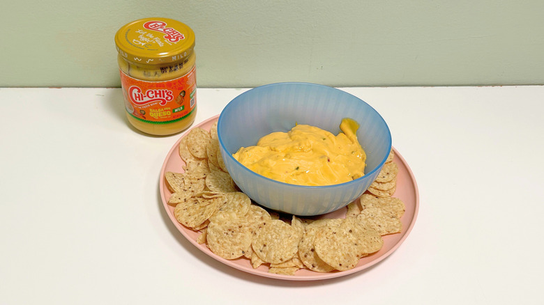 A bowl of queso surrounded by tortilla chips next to a jar of Chi-Chi's Mild Salsa Con Queso