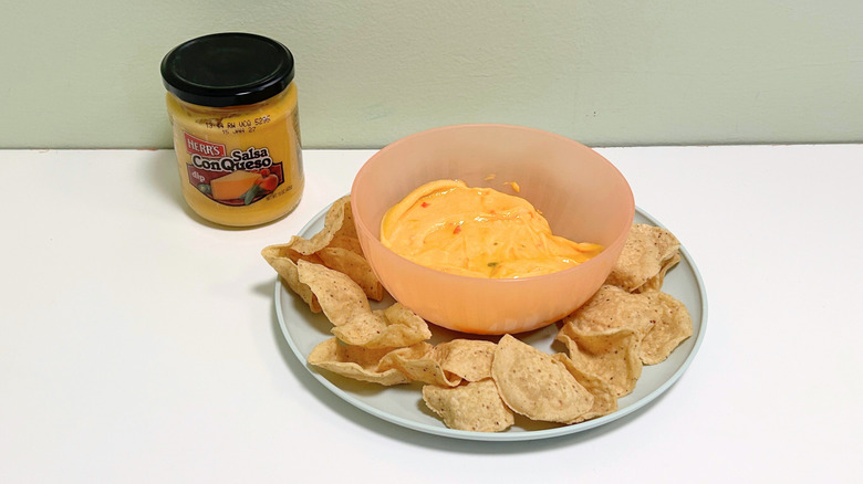 A bowl of queso surrounded by tortilla chips next to a jar of Herr's Salsa Con Queso