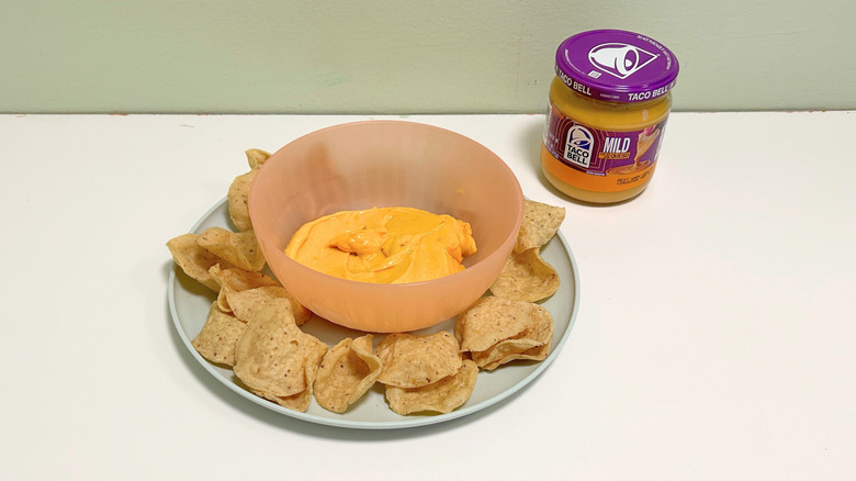 A bowl of queso surrounded by tortilla chips next to a jar of Taco Bell Mild Salsa Con Queso