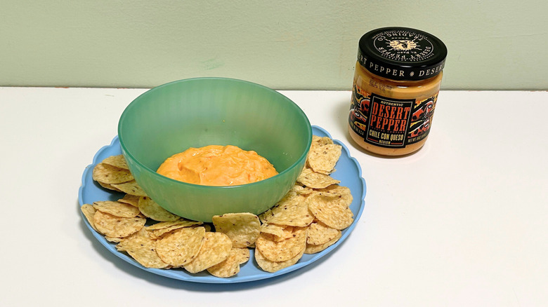 A bowl of queso surrounded by tortilla chips next to a jar of Desert Pepper Chile Con Queso