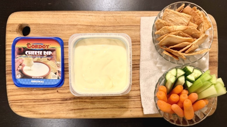 Gordo's Cheese Dip on a wooden board next to its lid, a bowl of pita chips, and a bowl of carrots, cucumbers, and celery sticks