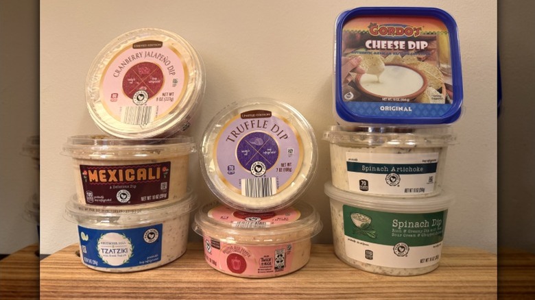 Aldi dips lined up against a wall, including Gordo's Cheese Dip, Tzatziki, Cranberry Jalapeno Dip, Roasted Red Pepper dip, Mexicali Dip, Truffle dip, Spinach Artichoke Dip, and Spinach Veggie dip