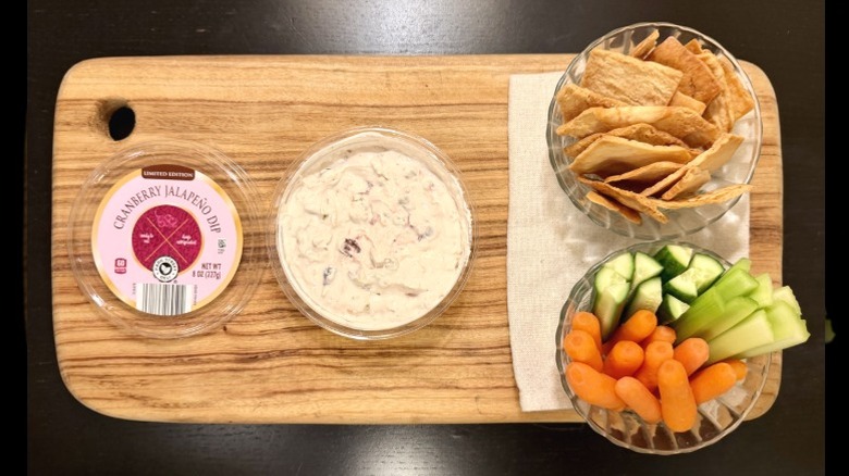 Park Street Deli Cranberry Jalapeño Dip on a wooden board next to its lid, a bowl of pita chips, and a bowl of carrots, cucumbers, and celery sticks