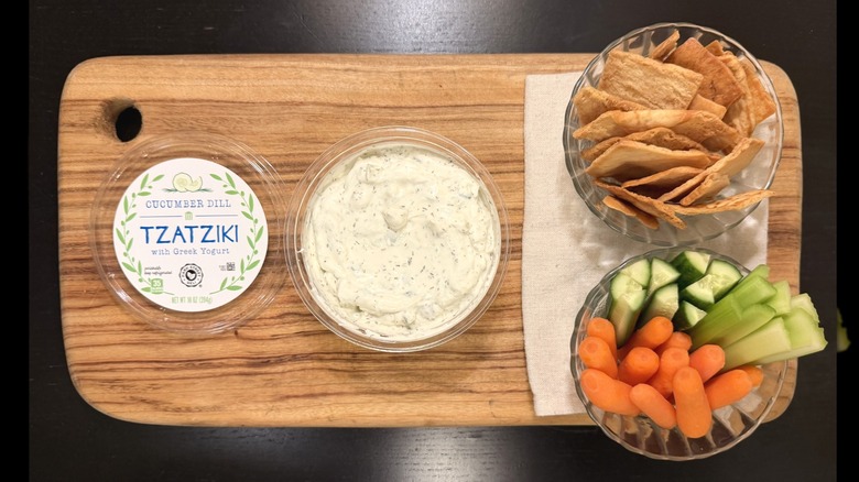 Park Street Deli Cucumber Dill Tzatziki on a wooden board next to its lid, a bowl of pita chips, and a bowl of carrots, cucumbers, and celery sticks