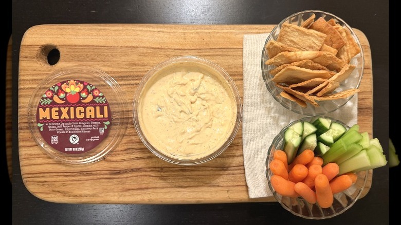 Park Street Deli Mexicali Dip on a wooden board next to its lid, a bowl of pita chips, and a bowl of carrots, cucumbers, and celery sticks