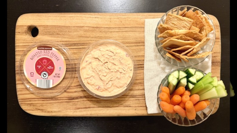 Park Street Deli Red Pepper Goat Cheese Dip on a wooden board next to its lid, a bowl of pita chips, and a bowl of carrots, cucumbers, and celery sticks