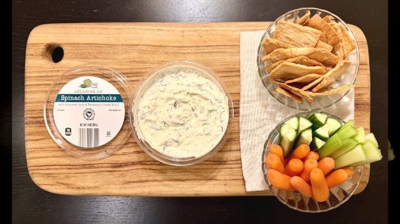 Park Street Deli Spinach Artichoke Dip on a wooden board next to its lid, a bowl of pita chips, and a bowl of carrots, cucumbers, and celery sticks