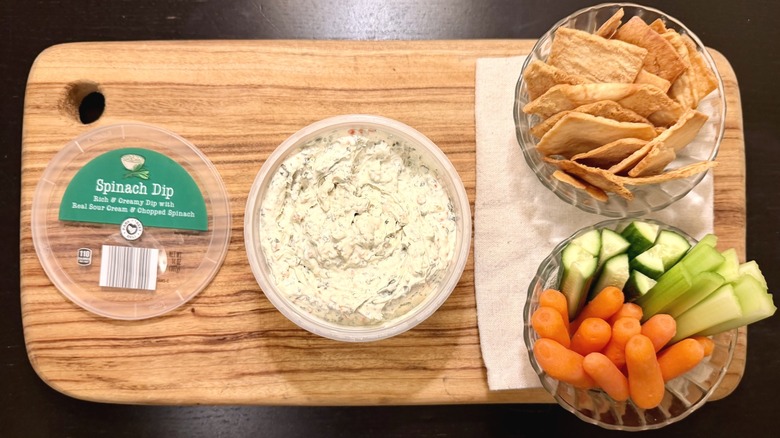 Park Street Deli Spinach Veggie Dip on a wooden board next to its lid, a bowl of pita chips, and a bowl of carrots, cucumbers, and celery sticks