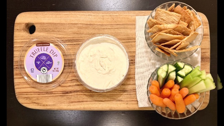 Park Street Deli Truffle Dip on a wooden board next to its lid, a bowl of pita chips, and a bowl of carrots, cucumbers, and celery sticks