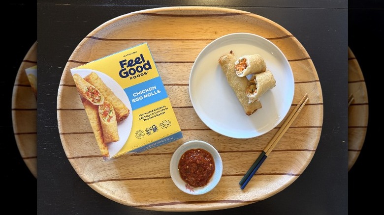 Feel Good Foods Gluten Free Chicken Egg Rolls on a wooden tray next to their box and a dish of sambal