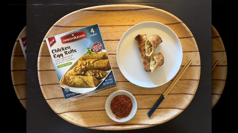 InnovAsian Chicken Egg Rolls on a wooden tray next to their box and a dish of sambal