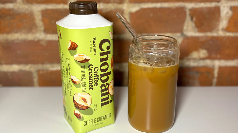 A yellow bottle of Chobani Hazelnut Coffee Creamer on a white table against a brick backdrop next to a glass cup of coffee