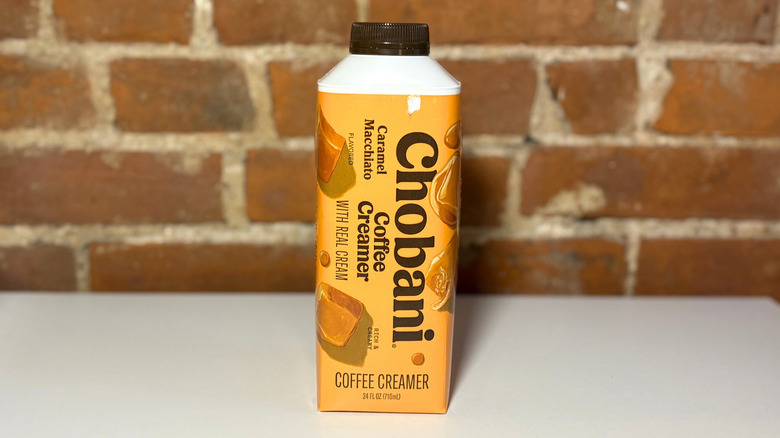 An orange bottle of Chobani Caramel Macchiato Coffee Creamer on a white table against a brick backdrop