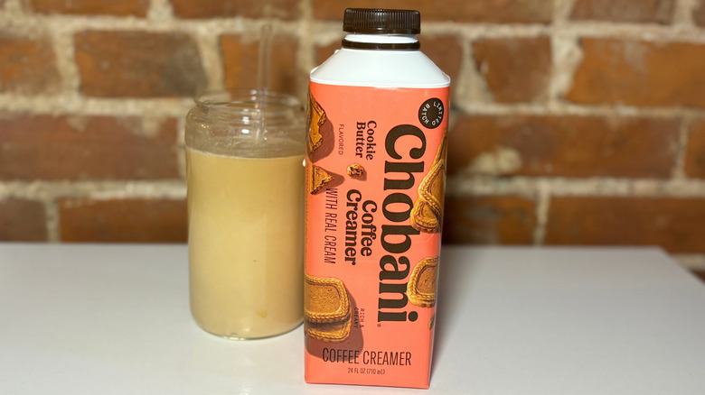 An orange bottle of Chobani Cookie Butter Coffee Creamer on a white table against a brick backdrop next to a glass of coffee