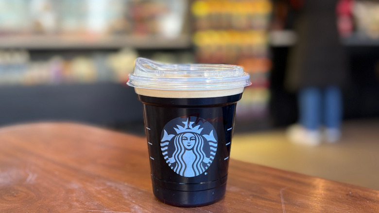 A black cup of a Starbucks drink on a wood table with a blurry background