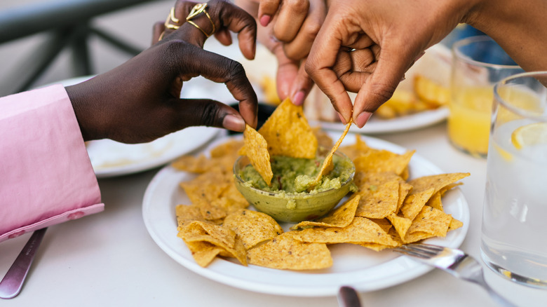 three hands reach over a plate of tortilla chips to dip chips in a bowl of guacamole