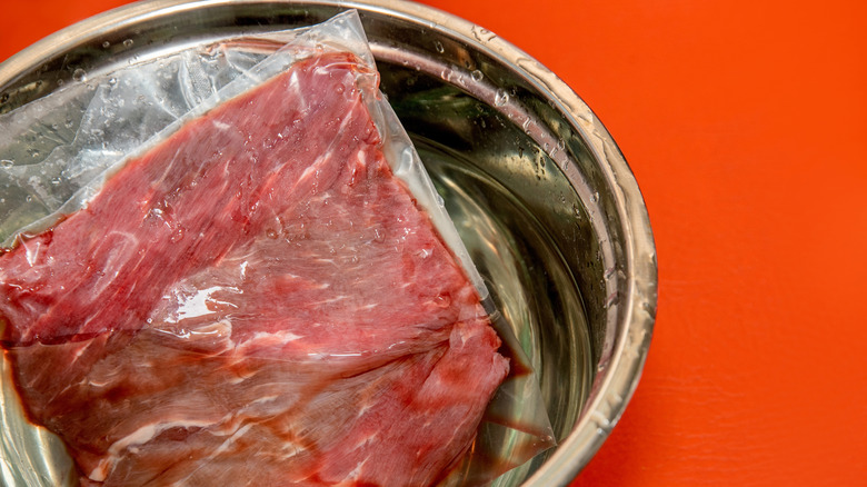 A raw beef cut thawing in its package in a metal bowl of water