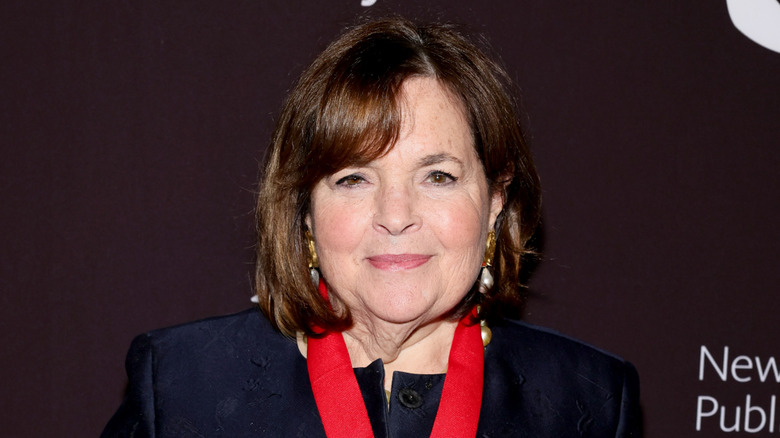 A close up of Ina Garten with a navy jacket and red lanyard against a plum background.