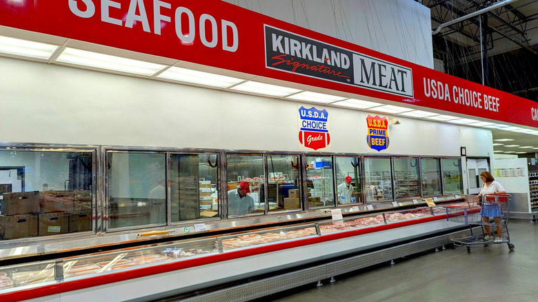 A woman with a shopping cart standing near the meat section of a Costco