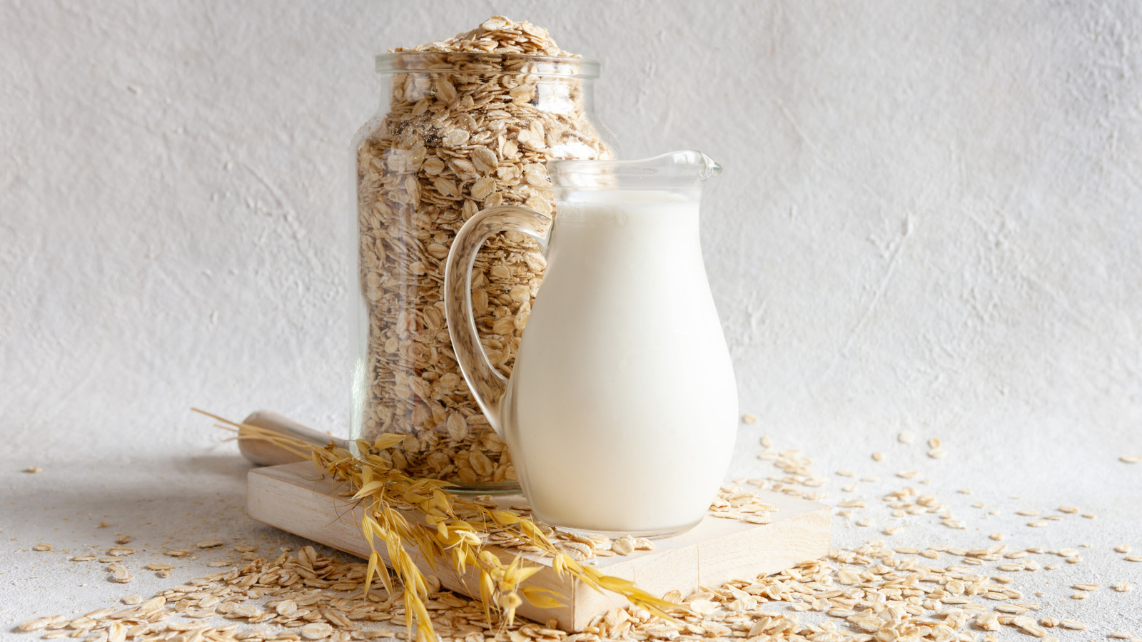 Jar of rolled oats next to a pitcher of creamy oat milk on a cutting board, illustrating oat milk and its oat source