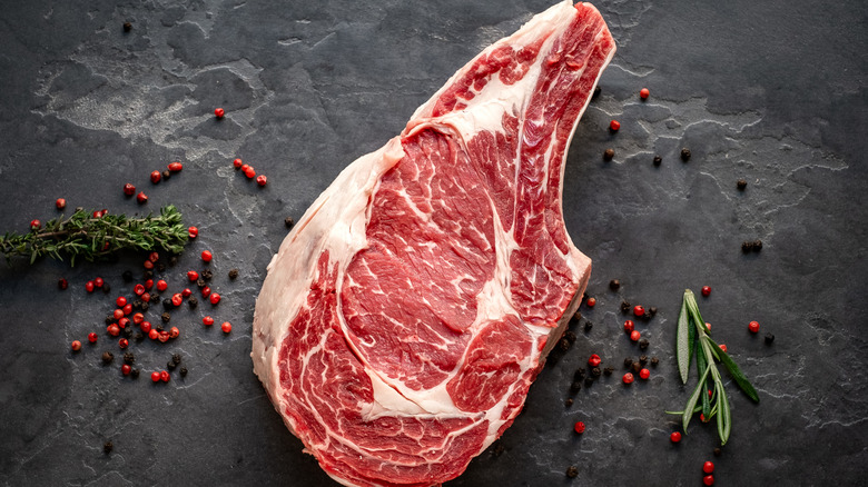 A raw cowboy steak on a slate slab with peppercorns and herbs