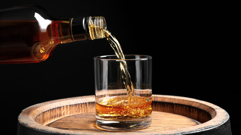 Whisky pouring from a bottle into a glass on top of a wooden barrel