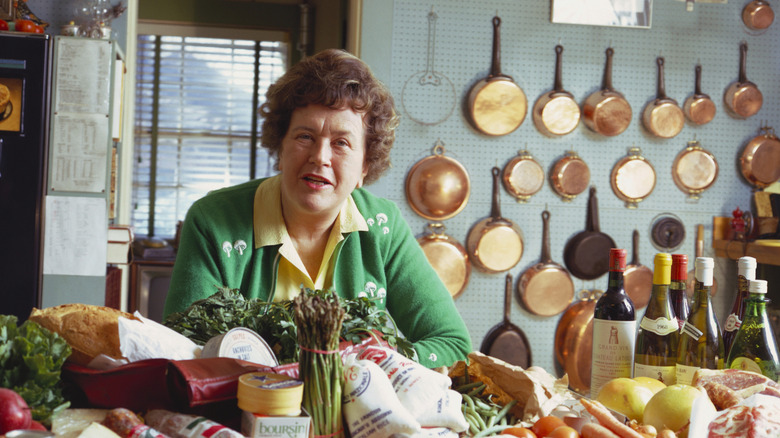 Julia Child in her kitchen, with copper pots behind her and ingredients in front of her