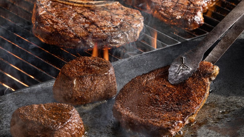 Variety of steaks on and next to the grill with one held by LongHorn-logoed tongs