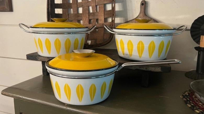 Three vintage white and yellow Cathrineholm enamel pots with lids and lotus leaves on shelves.