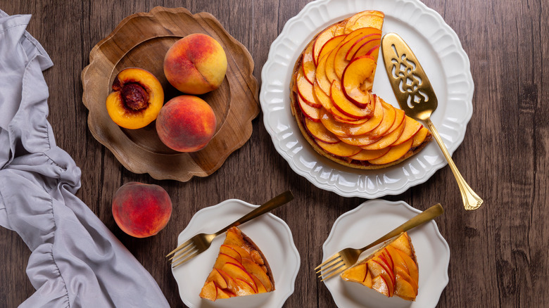 A peach tart on a white dish sits next to fresh peaches on a wooden dish