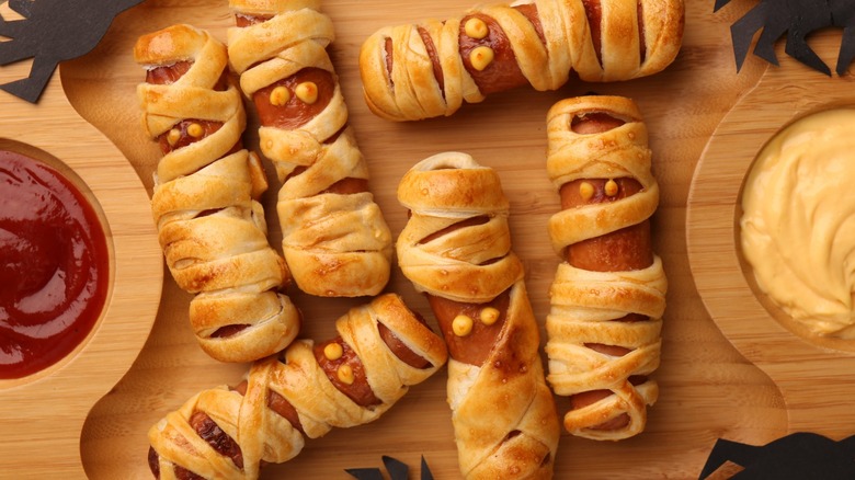 Mummy hot dogs on a cutting board with black cut-out spiders and ketchup and mustard