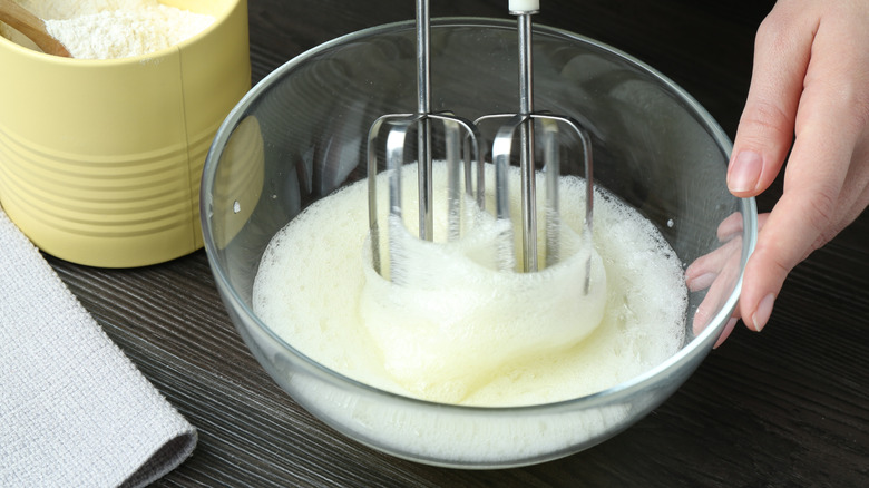 A person whisks egg whites in a clear bowl on a counter