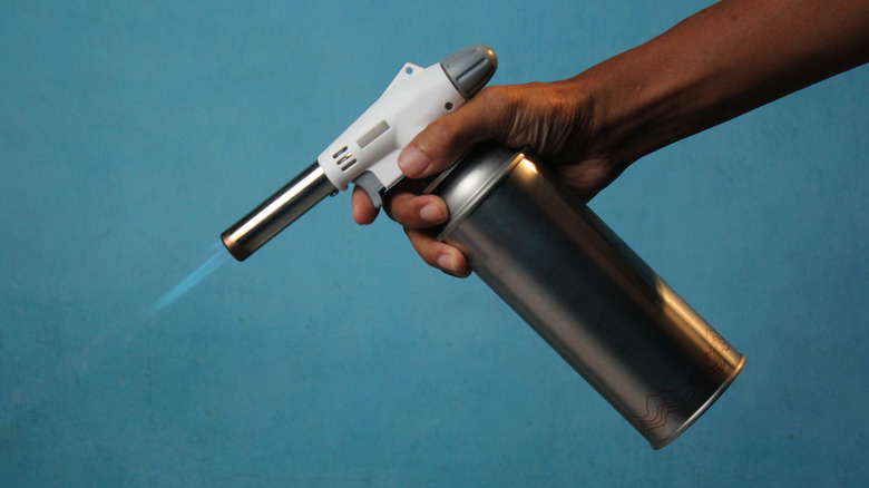 A hand holds a small butane-powered kitchen torch emitting a small blue flame