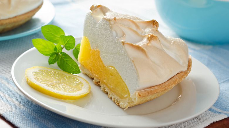 A piece of lemon meringue pie on a plate with a lemon slice and some greenery